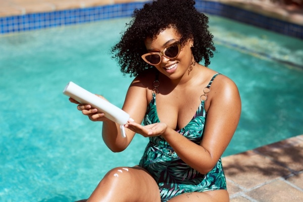 A woman in a green bathing suit sits beside a pool pouring sunscreen into her hand. Sunscreen is one of the best skincare tips. 