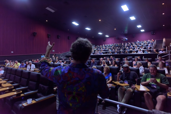 The silhouette of a man on a stage addressing a crowd of people sitting in movie theatre seats. 