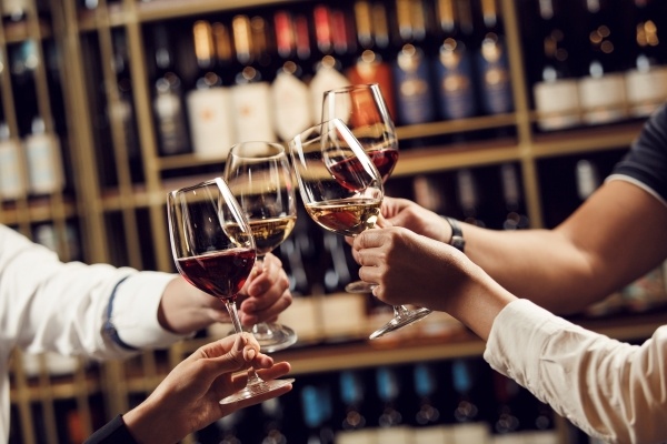 Four hands toast wine glasses with red and white wine in front of shelves of wine bottles.