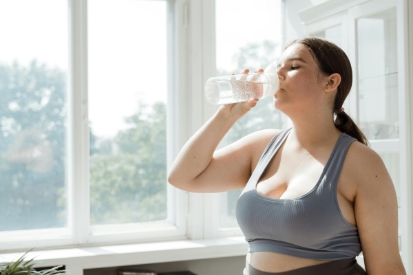 A woman in a gray sports bra standing by a window drinks water from a water bottle.