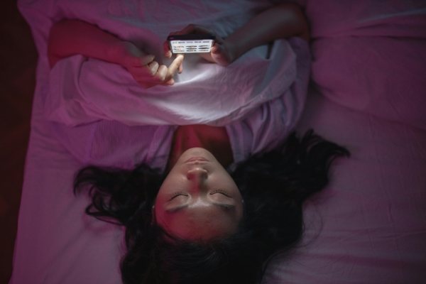 A woman lies on her back in bed in the dark, holding a glowing phone. 