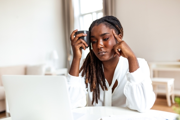 A tired-looking woman wearily sits at a desk and looks in a computer screen as she struggles with digital burnout.