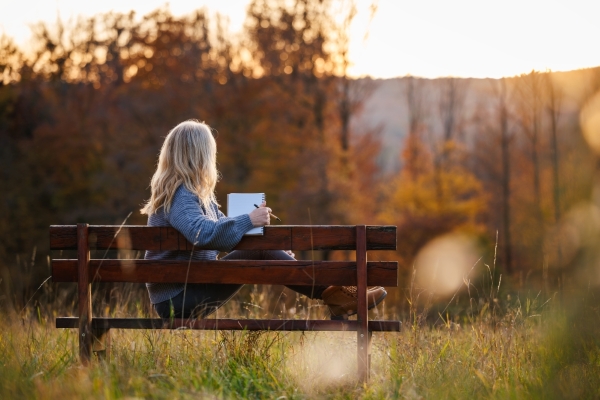 A woman sits on a bench in a grassy meadow holding a journal and looking out over trees at sunset.