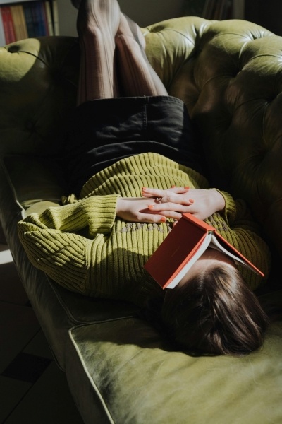 A woman lies on her back on a green couch with her feet in the air and a folded book resting on her face.