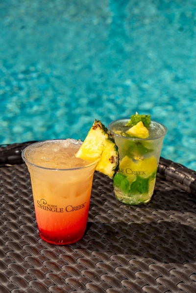 What Your Cocktail Order from Cat-Tails Pool Bar & Grille Says About You 7 A red ombre cocktail and a green and yellow cocktail on a wicker table by a sparkling blue pool in Rosen shingle Creek cups.