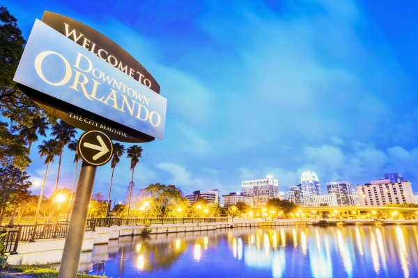 A sign reading Welcome to Downtown Orlando with a lake and the glowing Orlando skyline at twilight behind it. Orlando's MCO airport is soon to be home to many local restaurants. 