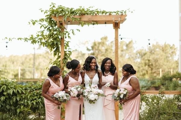 A bride and four bridesmaids in pale pink dresses stand under a wooden trellis draped with greenery.