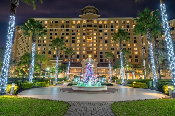 The exterior of Rosen Shingle Creek lit at night with a decorated Christmas tree, a glowing fountain, and palm trees wrapped in Christmas lights.
