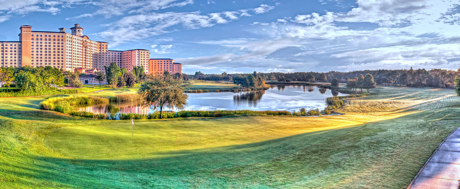 A beautiful hotel exterior towers against a blue cloudy sky in the background of a rolling golf course and a serene pond.