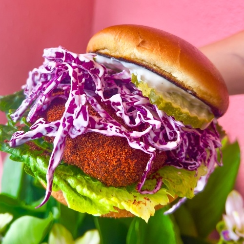 A close-up of a plant-based chicken sandwich with bright purple cabbage slaw and green lettuce in front of a pink background. From Dharma Southern Chick'n, a vegan dining establishment in Orlando. 