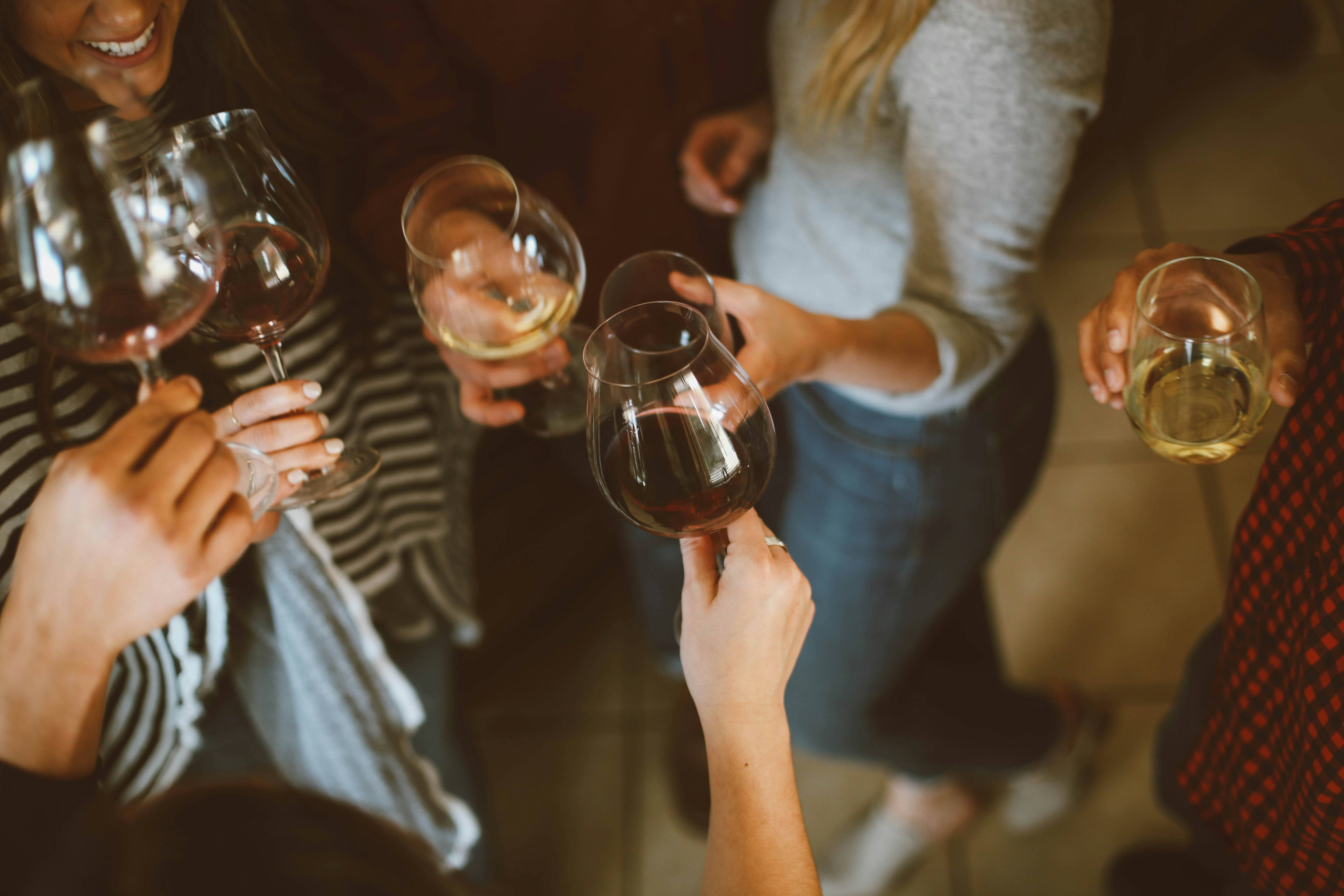 A dark, blurry image showing people (without their faces) holding wine glasses filled with different colored wine in each.