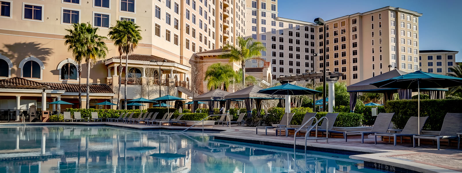 A pool surrounded by lounge chairs, blue umbrellas, palm trees, and a beautiful hotel in the background.