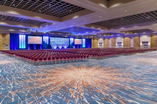 A large ballroom with an orange sunburst patterned carpet, hundreds of chairs in rows, and a stage with multiple screens. 