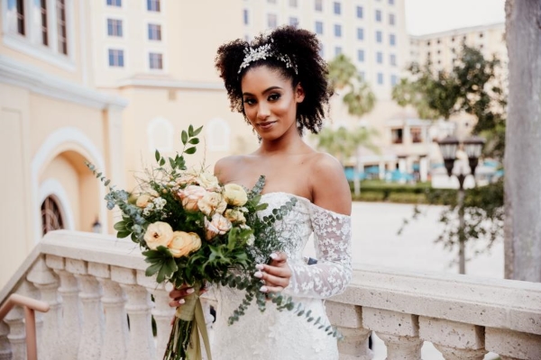 A bride in an off-shoulder wedding gown holds a peach colored bouquet standing on a stone balcony overlooking Rosen Shingle Creek.