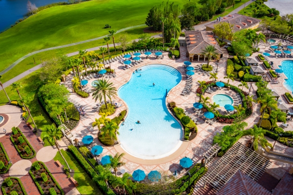 An aerial view of a curved swimming pool surrounded by palm trees, blue umbrellas, and a rolling golf course.