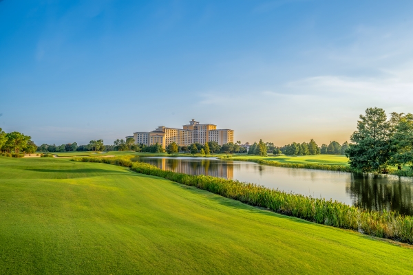 A stretch of green lawn surrounding a beautiful lake with a large hotel towering in the distance during twilight. 