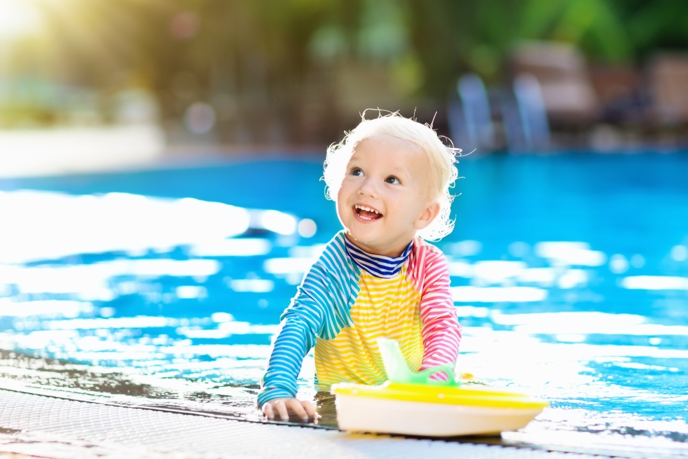 A young child in long-sleeved protective swimwear smiles, sitting beside a pool. 