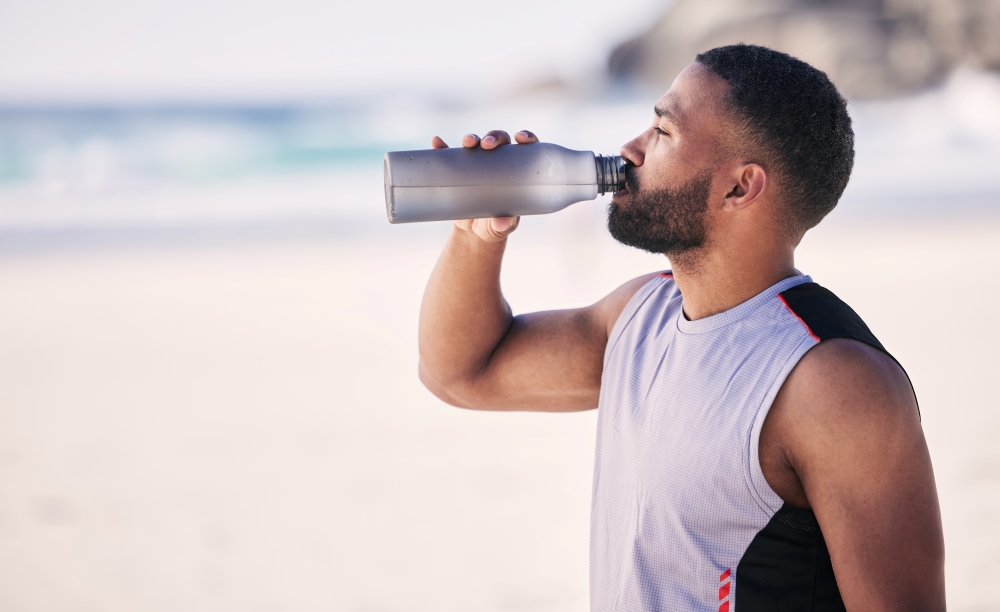 A man in a sleeveless shirt drinks from a water bottle on a beach. 
