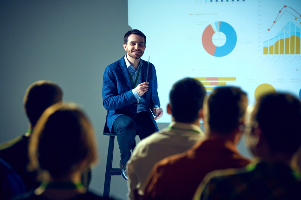 A man in a blue suit sits in front of a projection screen, addressing a crowd of silhouetted people.