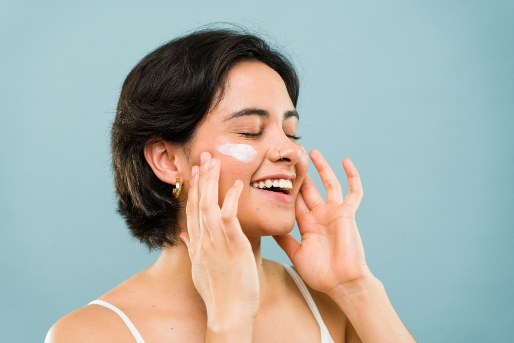 A woman rubs moisturizer onto her skin, smiling. 