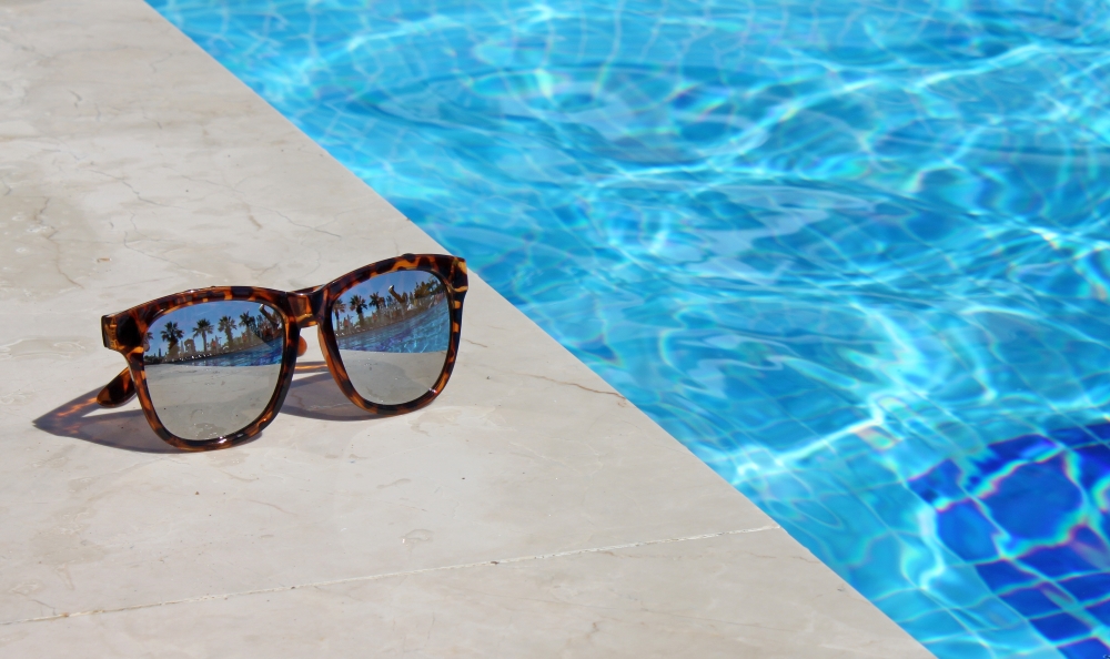 A pair of sunglasses sits on a pool deck beside sparkling blue pool water. 