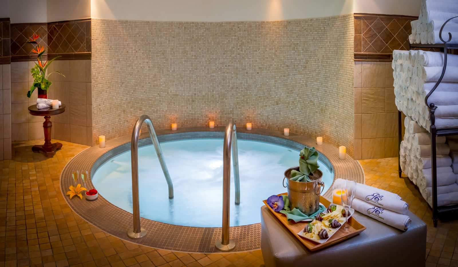 A blue whirlpool with metal railings in a dimly lit tiled room with candles, a towel rack, and a plate of chocolate covered strawberries and Champagne bucket