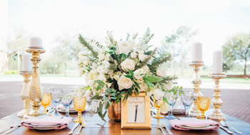 A wedding table setting with gold candle pillars, a white centerpiece with greenery, plates, glassware, and a framed table number 