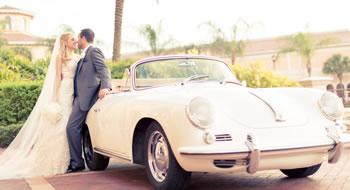 A bride and groom lean on a white vintage convertible with palm trees in the background.
